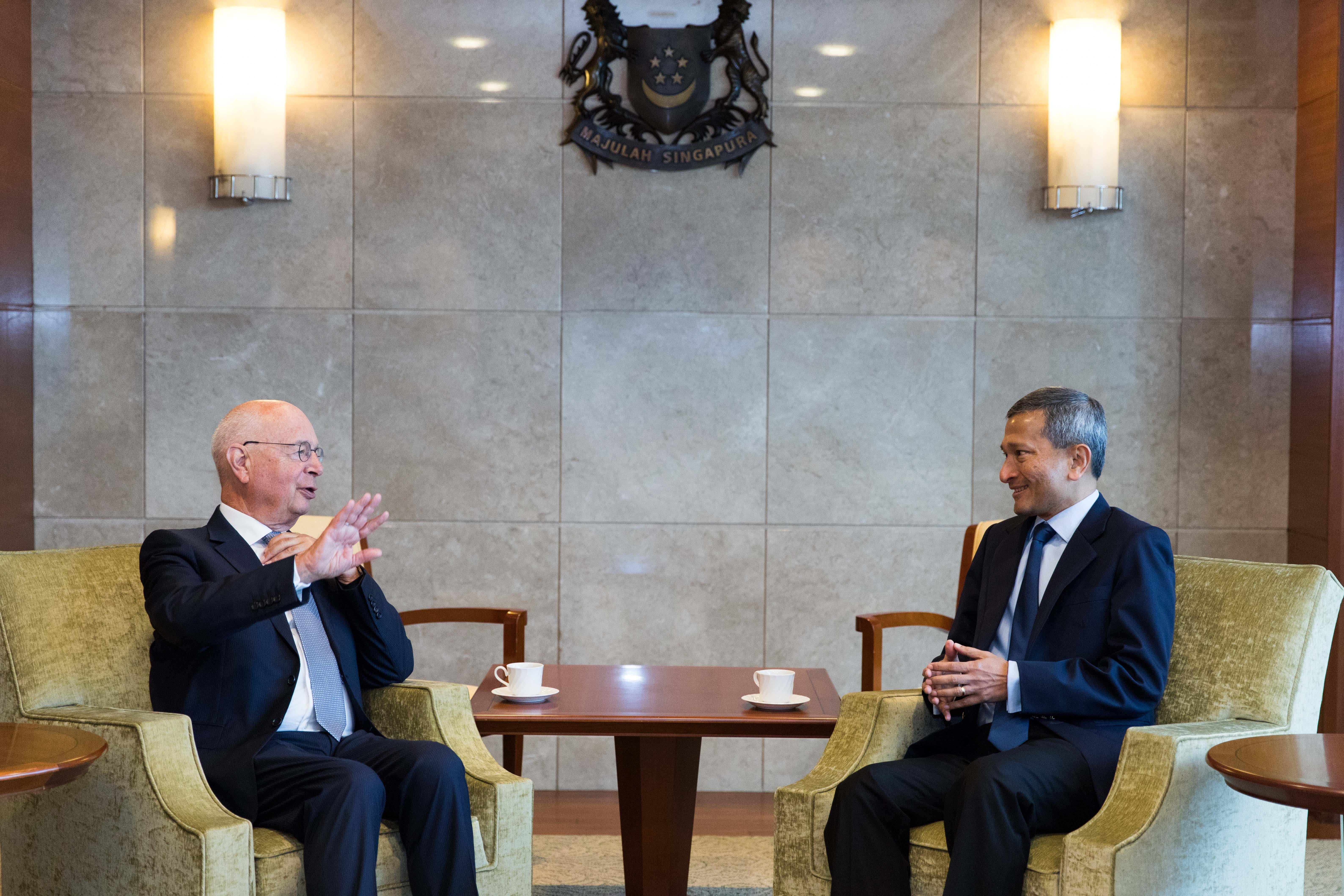 Klaus Schwab and Lee Hsien Loong in suits seated, having a discussion in front of the Singapore crest.